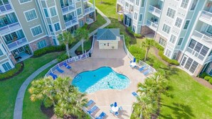 An aerial view of a residential building complex with a central swimming pool surrounded by lounge chairs and palm trees. - Amazing Pond View condo + pool at MyrtleWoods (Myrtle Beach)