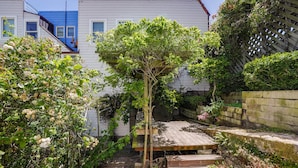 Un étroit sentier mène à une terrasse en bois surmontée d’une pergola, le tout étant entouré d’une végétation luxuriante et d’un bâtiment blanc doté d’un toit bleu.