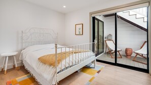 Une chambre avec un lit en métal blanc, une petite table de chevet blanche, un tapis coloré et une porte-fenêtre coulissante donnant sur un balcon équipé de deux chaises blanches et d’un pot de fleurs.