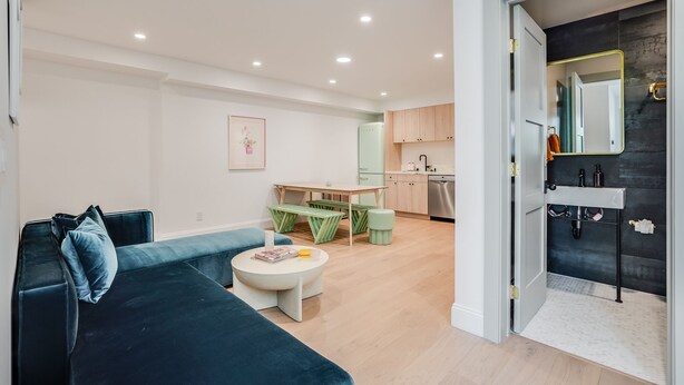 Un salon moderne avec un canapé bleu foncé, une table basse ronde blanche et un ensemble de salle à manger vert. Une cuisine avec des meubles en bois et un plan de travail noir est visible à travers une porte ouverte.