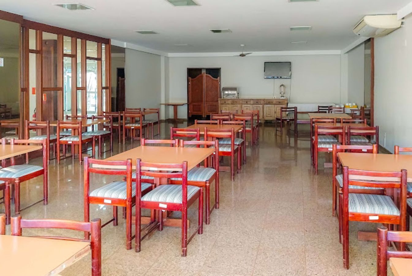 Un comedor con varias mesas y sillas de madera, una zona de cocina con armarios, y un televisor montado en la pared.