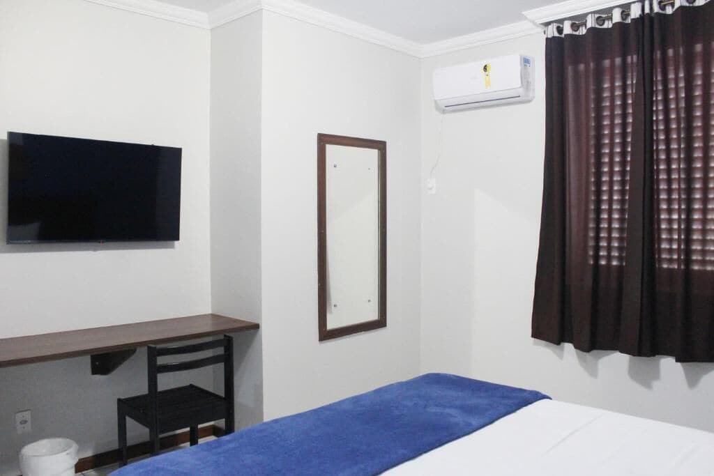 Quarto de hotel com cama, escrivaninha, cadeira, televisão, espelho e janela com cortinas.