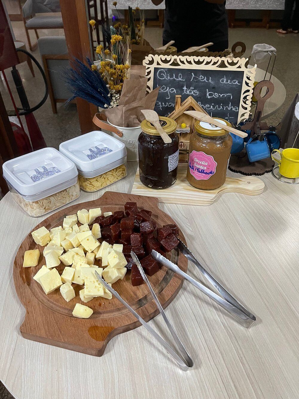 A wooden cutting board with cheese and meat, jars of preserves, and a chalkboard with writing.