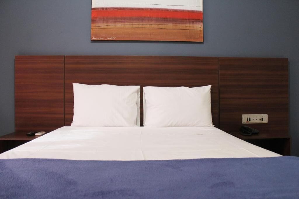 A hotel room with a wooden headboard, a bed with white linens, a blue blanket, and a painting on the wall.