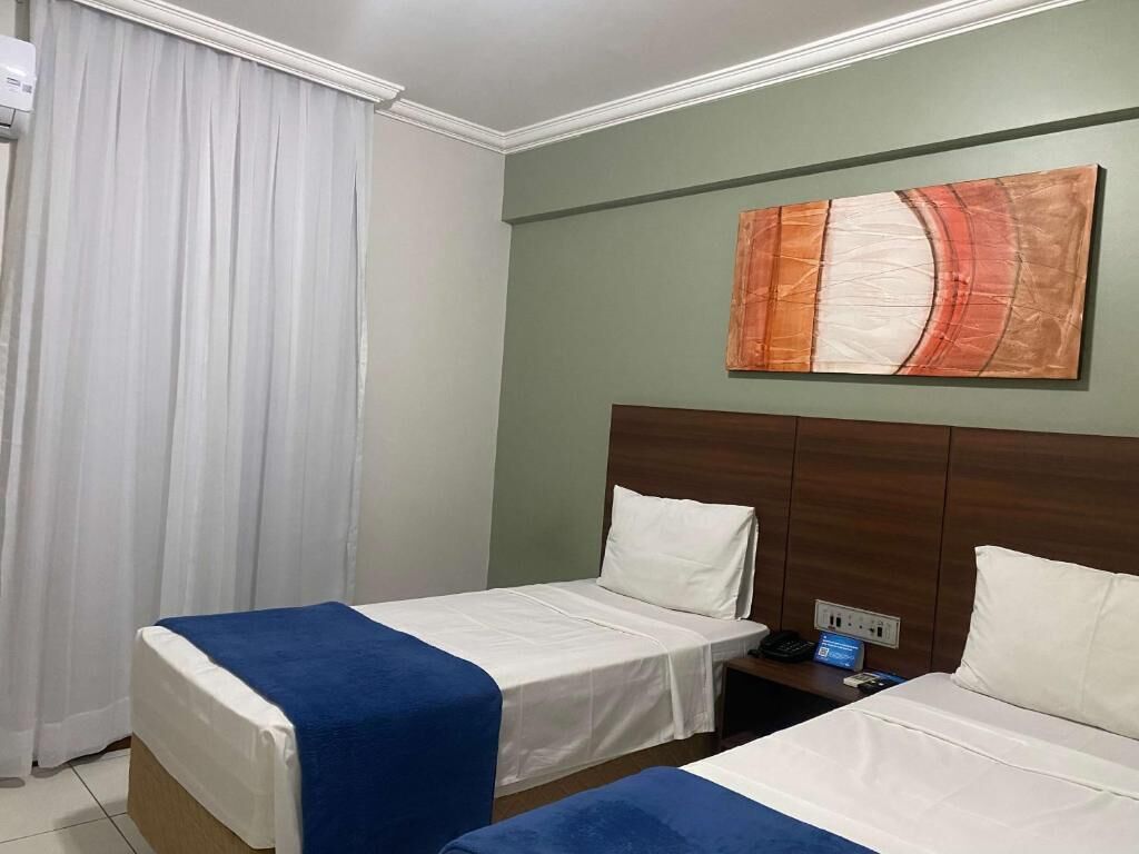 A hotel room with two single beds, a nightstand, a wall-mounted painting, and a window with curtains.