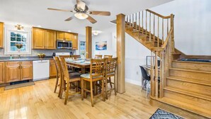 Cocina amplia con armarios de madera, una mesa de comedor y sillas. Se observa una escalera de madera con barandilla.