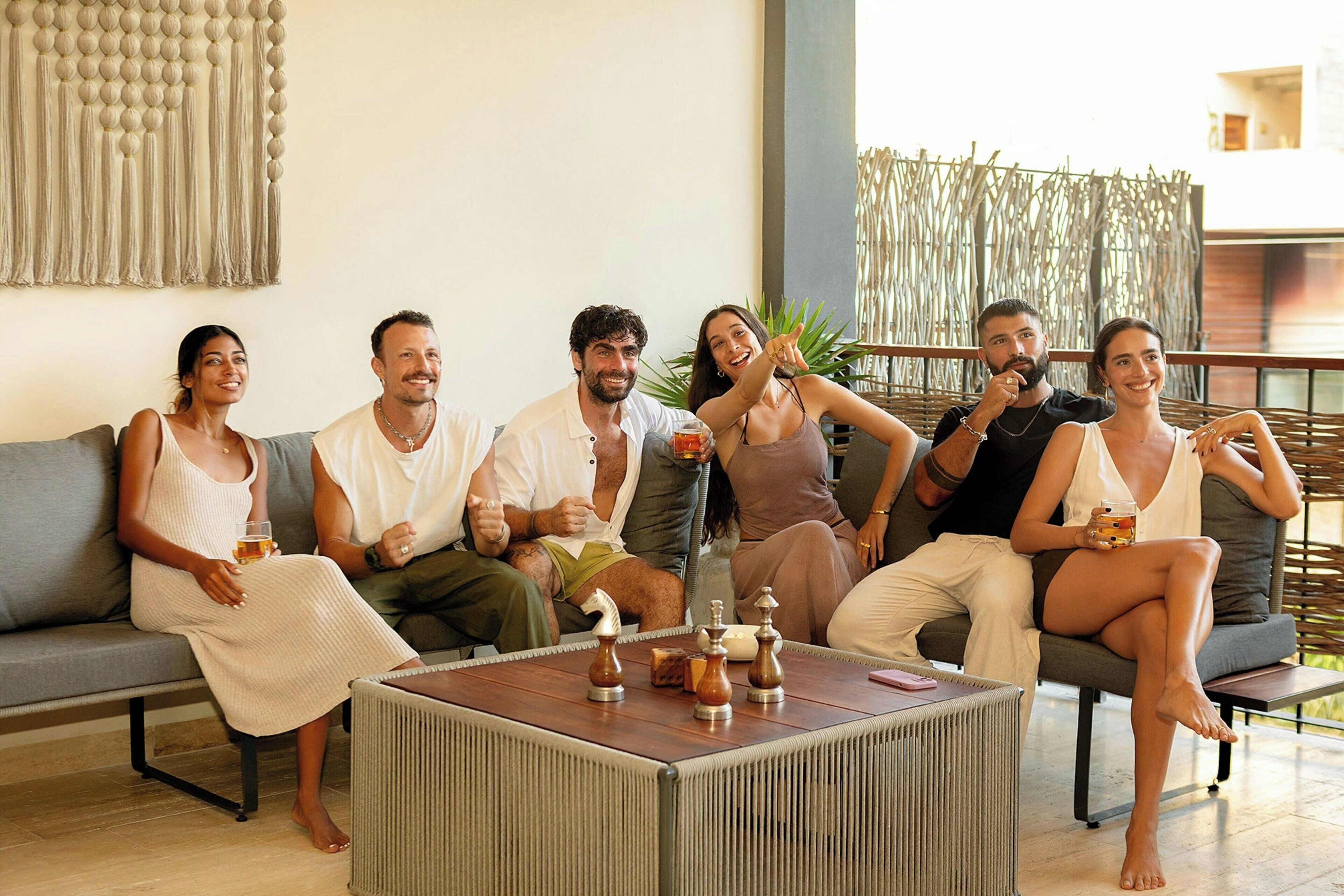 Un groupe de personnes est assis sur un canapé gris dans une pièce bien éclairée, au décor moderne. On y trouve une table basse en bois ornée d’objets décoratifs et une grande tapisserie murale tissée.