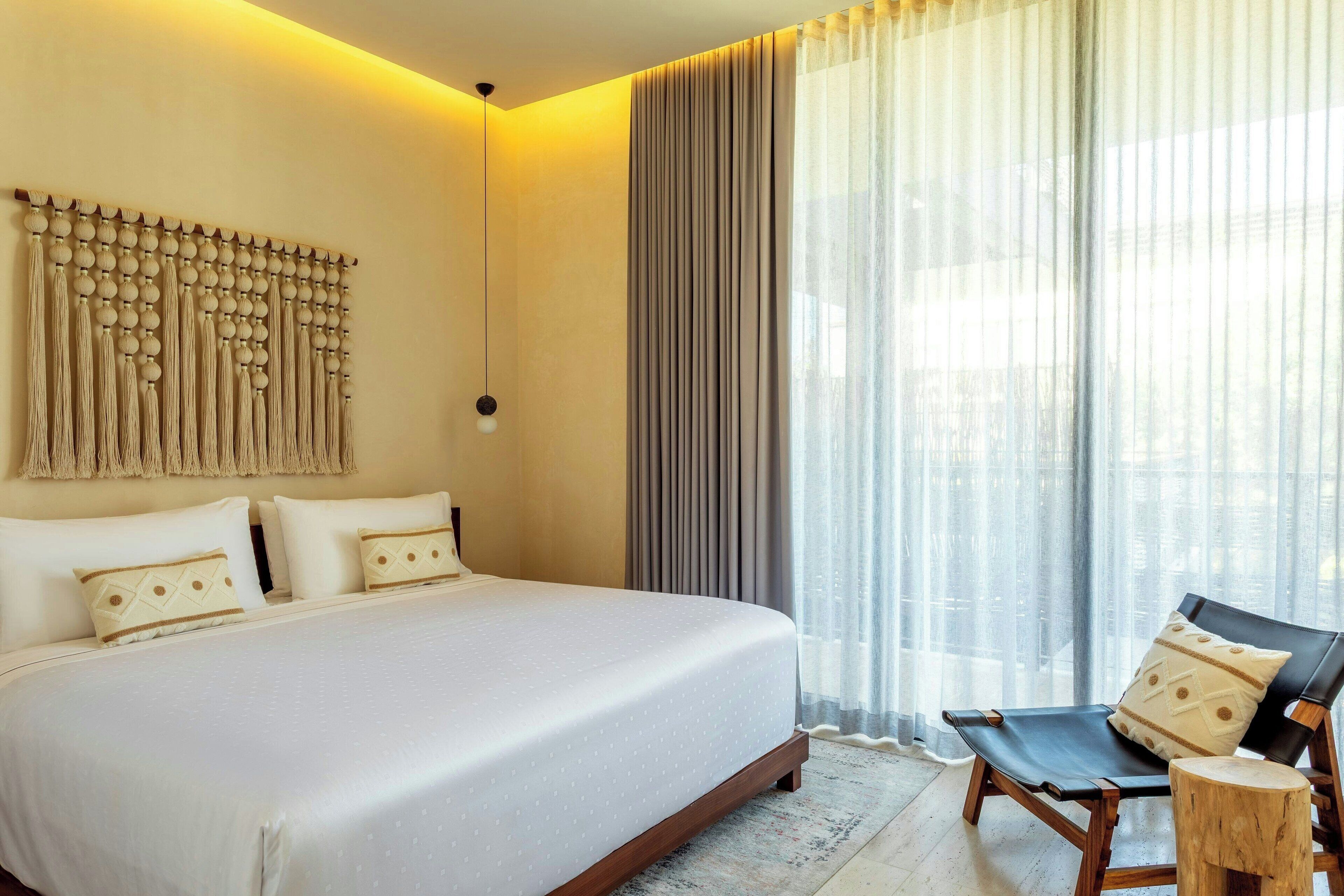 A hotel room with a large bed, a chaise lounge, a wooden headboard, and a view through sheer curtains.