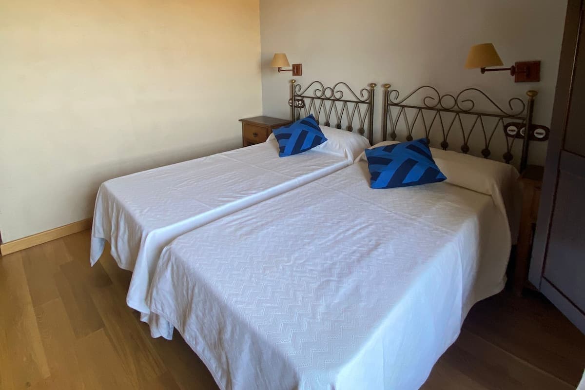 Habitación con dos camas individuales, suelo de madera, techo inclinado con vigas a la vista y dos lámparas de noche.