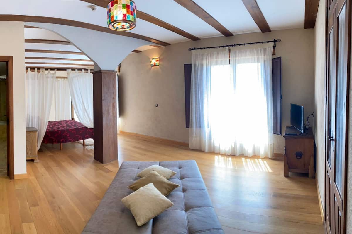 Amplia sala de estar con piso de madera, sofá gris, escritorio de madera y un gran ventanal con cortinas.