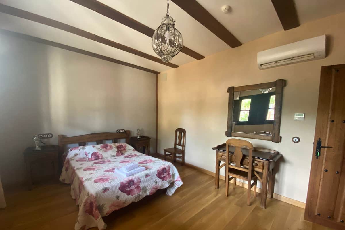 Un dormitorio con cama de madera, un escritorio y una silla. Se observa una araña, un equipo de aire acondicionado y una puerta.