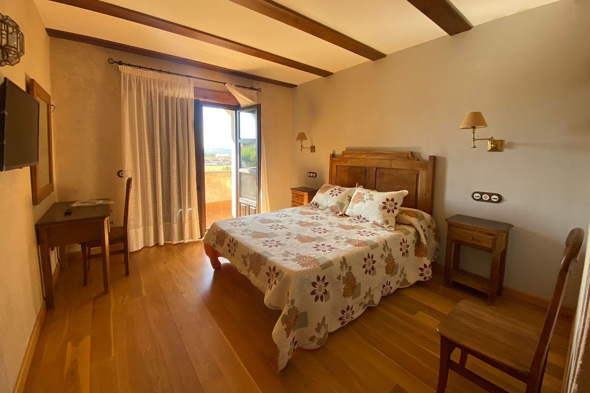 Un dormitorio con cama de madera, un escritorio, una silla, una mesita de noche y una lámpara. Hay una ventana con cortinas y vistas al exterior.
