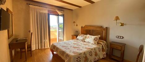 Un dormitorio con cama de madera, un escritorio, una silla, una mesita de noche y una lámpara. Hay una ventana con cortinas y vistas al exterior.