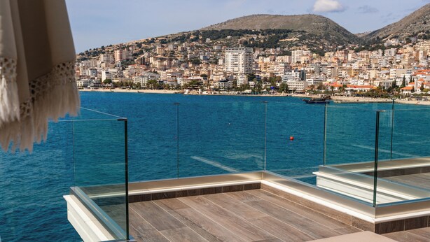 A terrace with a view of a coastal city, featuring a pool, seating area, and a glass railing. - Demi Hotel (Sarandë)