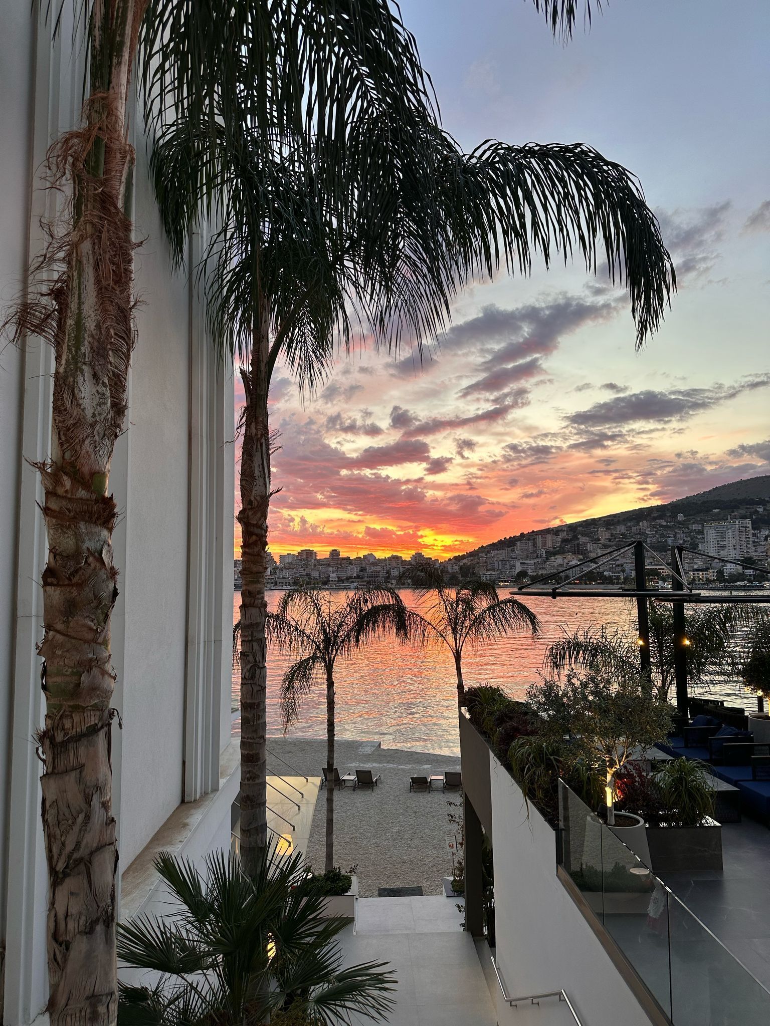 A balcony with a view of a sunset over a body of water, palm trees, and a cityscape in the background.
