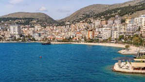 A coastal cityscape with clear blue waters, a sandy beach with lounge chairs, and a backdrop of residential buildings and hills. - Demi Hotel (Sarandë)