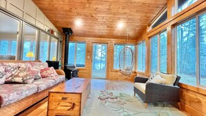 A cozy cabin interior with wooden walls, large windows, a sofa, a chair, and a hanging chair. - Hot Tub in the Forest Cozy Home on Peaceful Lake (Mountain)