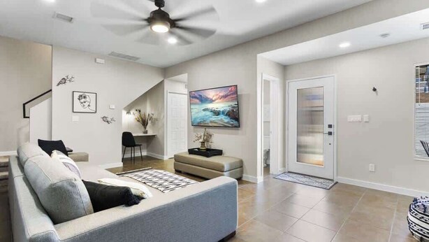 A modern living room with a sofa, a ceiling fan, a flat-screen TV, and a door with a glass panel. - Charming 4BR Near Fort Bliss (El Paso)