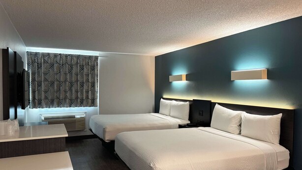 A hotel room with two beds, a desk, a window with patterned curtains, and a dark accent wall with lighting. - Studio 6 Carson, CA (Carson)