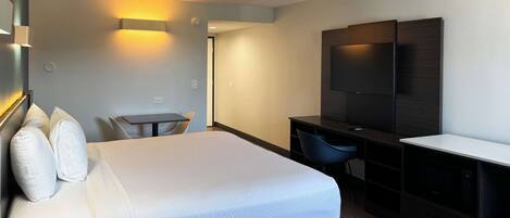 A hotel room with a bed, a desk, a chair, a TV, and a wall-mounted light.