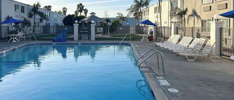 Ein Swimmingpool mit Liegestühlen und Sonnenschirmen, umgeben von Gebäuden und Palmen.