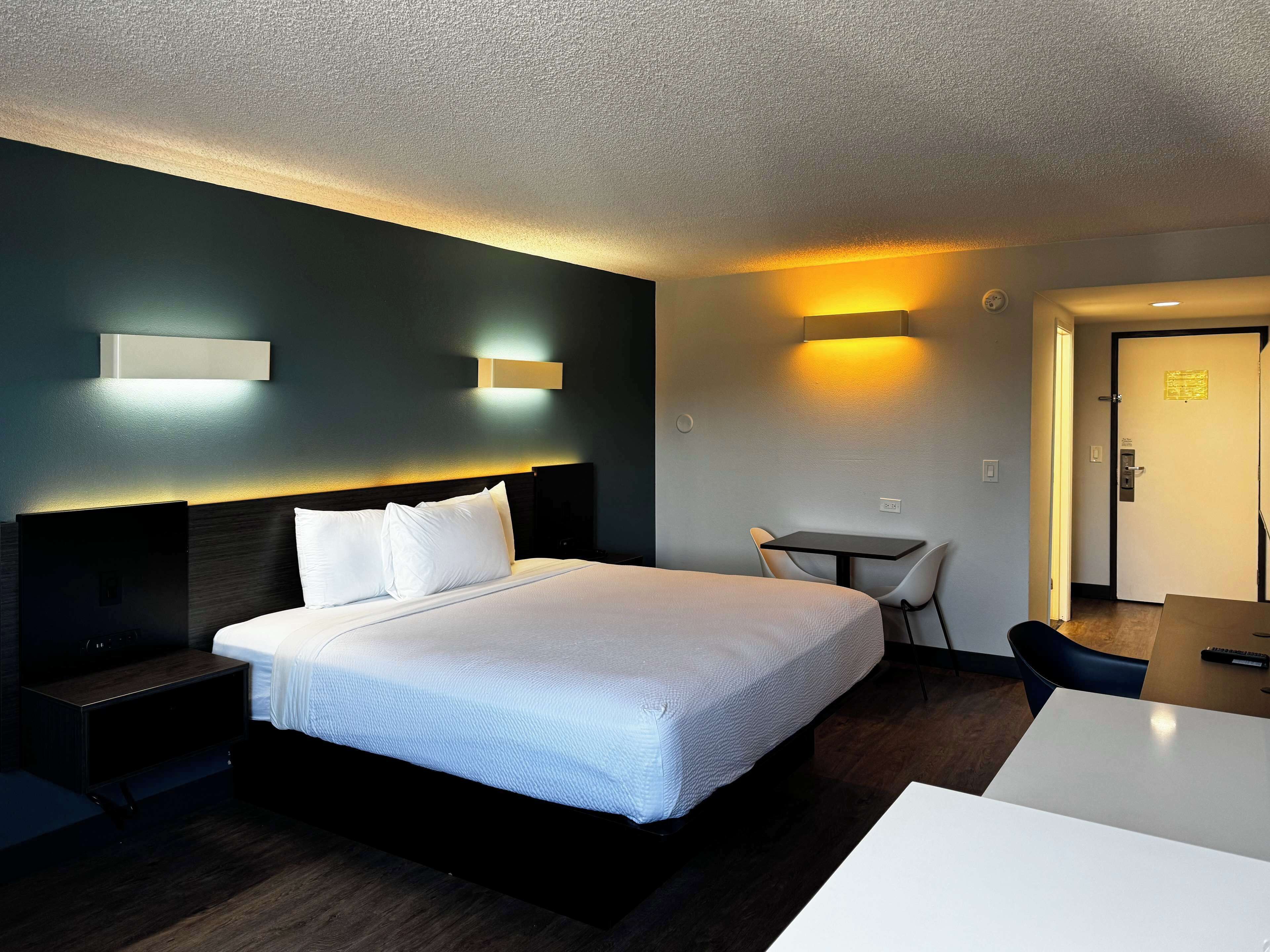Une chambre d’hôtel avec un grand lit, des tables de chevet, un bureau et une chaise. Il y a des lampes fixées au mur et une porte avec une pancarte.