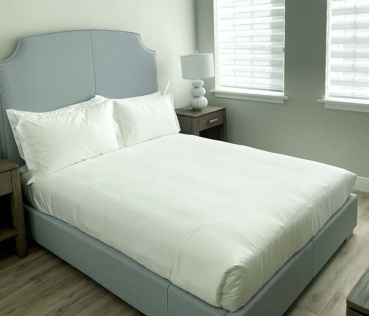 Een netjes opgemaakt bed met een gestreept hoofdeinde en voeteinde, een nachtkastje met een lamp, en een raam met jaloezieën.