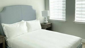 A neatly made bed with a striped headboard and footboard, a bedside table with a lamp, and a window with blinds. - The Pelican Villa (Nassau)