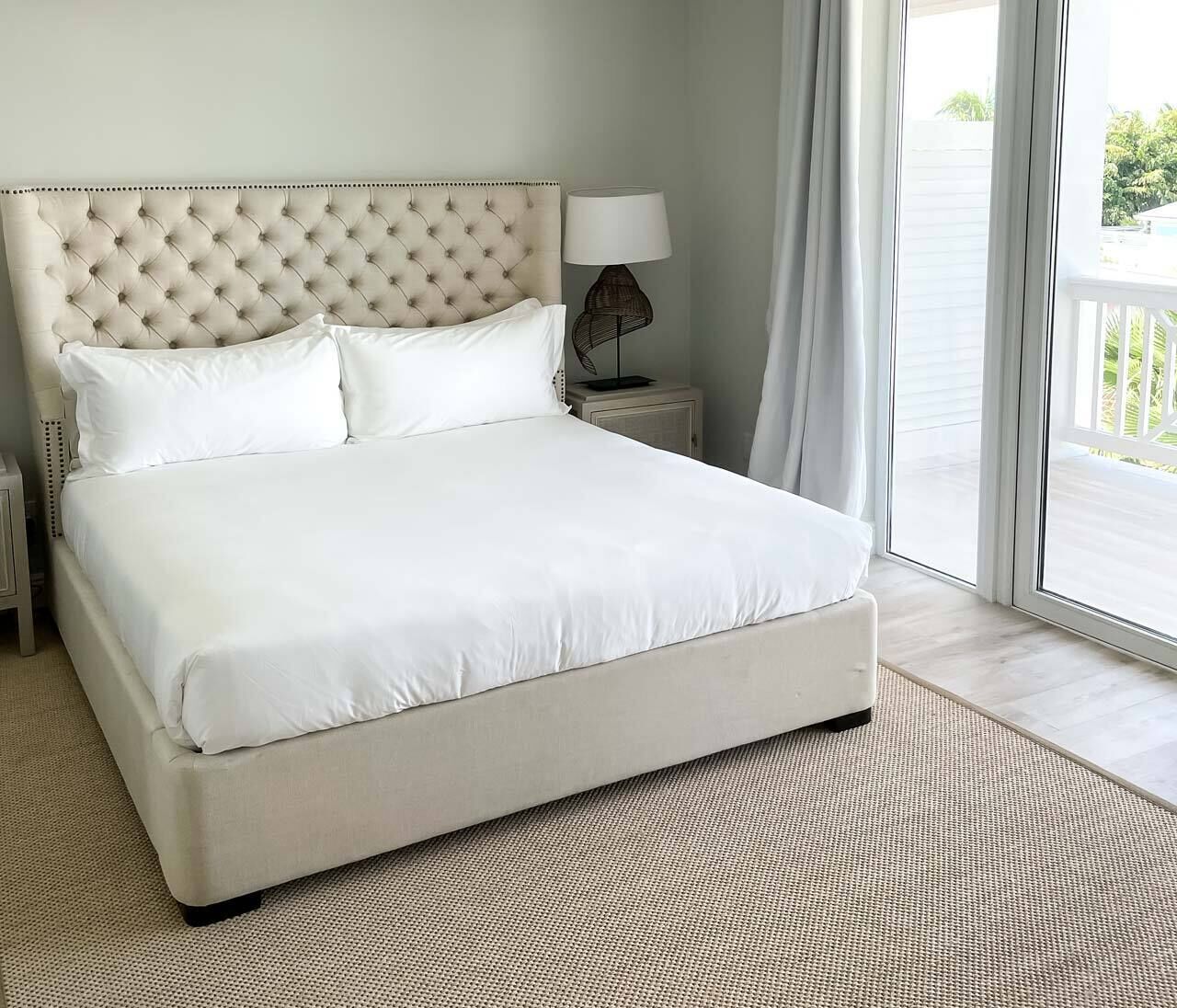 A large bed with a tufted headboard, white bedding, a small bedside table with a lamp, and a view of the outdoors through a sliding door.