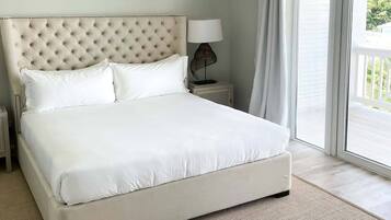A large bed with a tufted headboard, white bedding, a small bedside table with a lamp, and a view of the outdoors through a sliding door.
