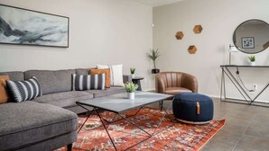 Una sala moderna con un sofá gris, una mesa de centro, un sillón de cuero marrón, un espejo redondo y una alfombra estampada.