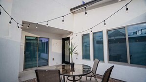 Mesa de vidrio con sillas, luces colgantes y una planta en maceta en un espacio exterior cubierto con ventanas.