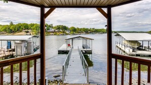 A wooden dock leading to a covered boat launch area with a view of a calm lake and houses in the background. - Lakefront Home w/ Private Dock in Sunrise Beach! (Sunrise Beach)