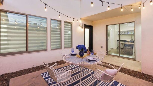 Um pátio externo com uma mesa redonda, cadeiras, luzes de festa e vista para a cozinha através de uma porta de vidro.