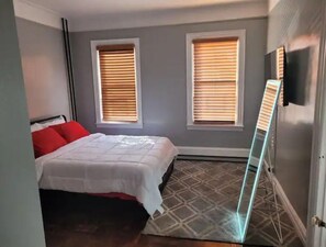 Ein Schlafzimmer mit einem Bett, zwei Fenstern mit Jalousien, einem an der Wand montierten Fernseher und einem gemusterten Teppich.