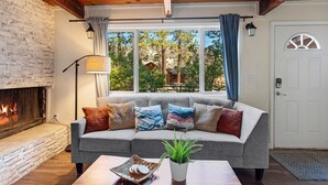 Una sala de estar acogedora con chimenea, un sofá gris con cojines de colores, techo de madera y vistas a árboles a través de amplias ventanas.