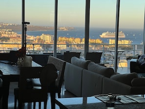 A modern living room with a large window overlooking a cityscape and the sea, featuring a sofa, dining table, and a telescope.
