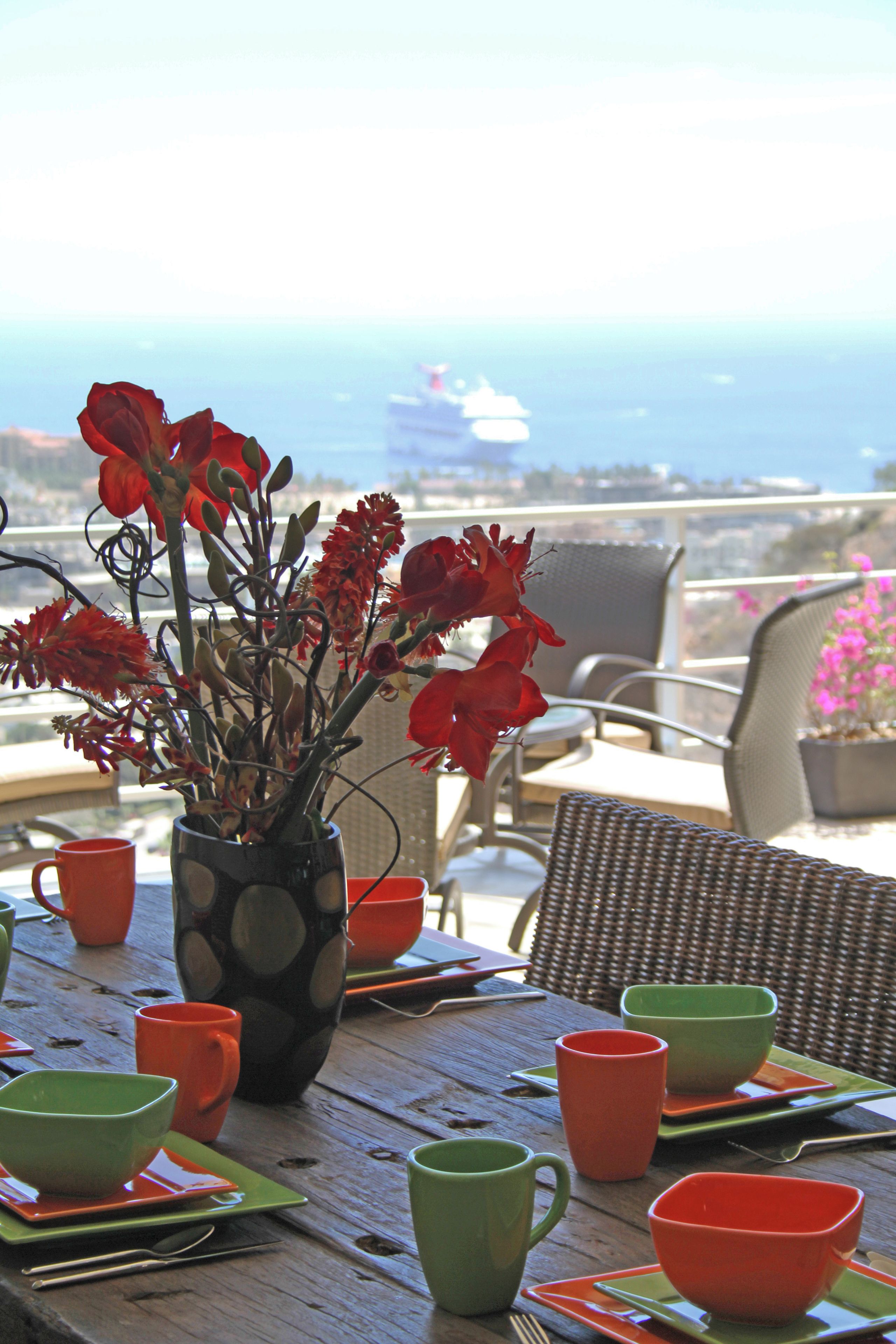 A table with a vase of red flowers, green and orange cups, and a view of the ocean and a cruise ship in the distance.