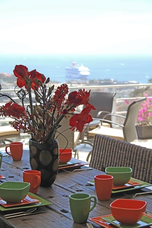 A table with a vase of red flowers, green and orange cups, and a view of the ocean and a cruise ship in the distance.