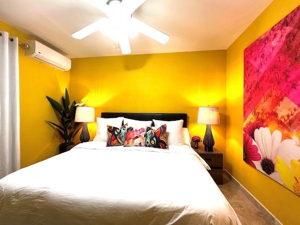 A hotel room with a bed, two bedside tables, a ceiling fan, an air conditioning unit, and a large floral wall mural.
