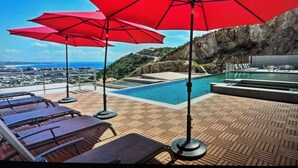 Uma área à beira da piscina com guarda-sóis vermelhos, espreguiçadeiras e vista para a costa.