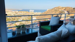 Varanda com móveis de vime, mesa de vidro e vista para a paisagem urbana costeira.