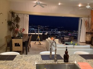 A modern living room with a city view, a dining area, and a kitchen with a sink.