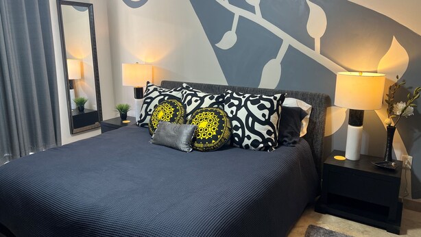 Uma cama com colcha azul escura, várias almofadas estampadas, duas luminárias de cabeceira, um espelho grande e uma parede com design decorativo.