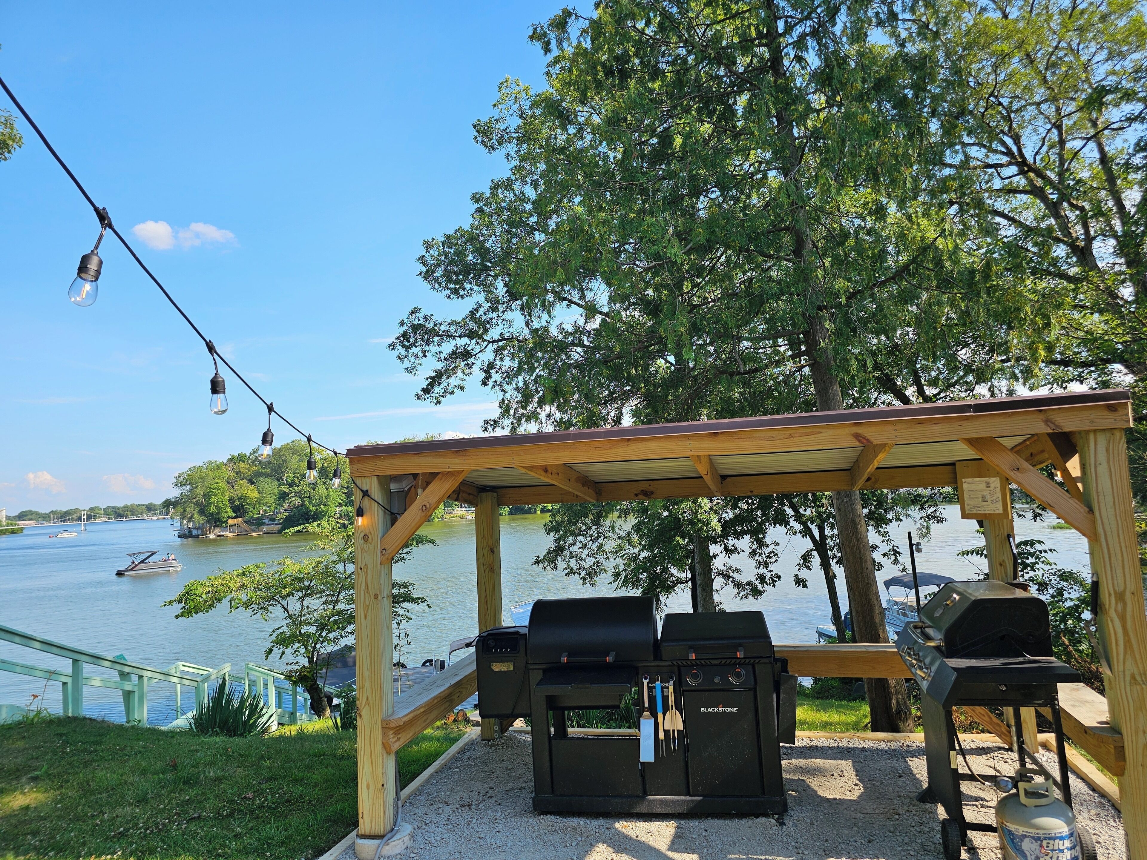 A wooden gazebo with barbecue grills, a grill lighter, and a propane tank, overlooking a lake with boats and a bridge in the background.
