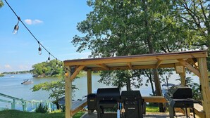 A wooden gazebo with barbecue grills, a grill lighter, and a propane tank, overlooking a lake with boats and a bridge in the background. - Key Lime Resorts - Unit #1 RV Retreat Villa (Monticello)