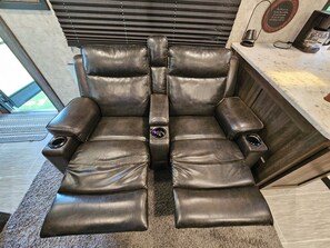 Un sillón de cuero reclinable con portavasos y panel de control, ubicado en una habitación con ventana y persianas.