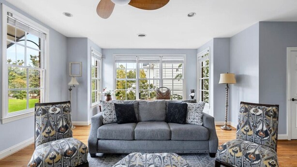 A living room with a grey sofa, patterned armchairs, a ceiling fan, and large windows with a view of greenery. - Lake Carolina Condo | 3BR 2.5BA (Columbia)