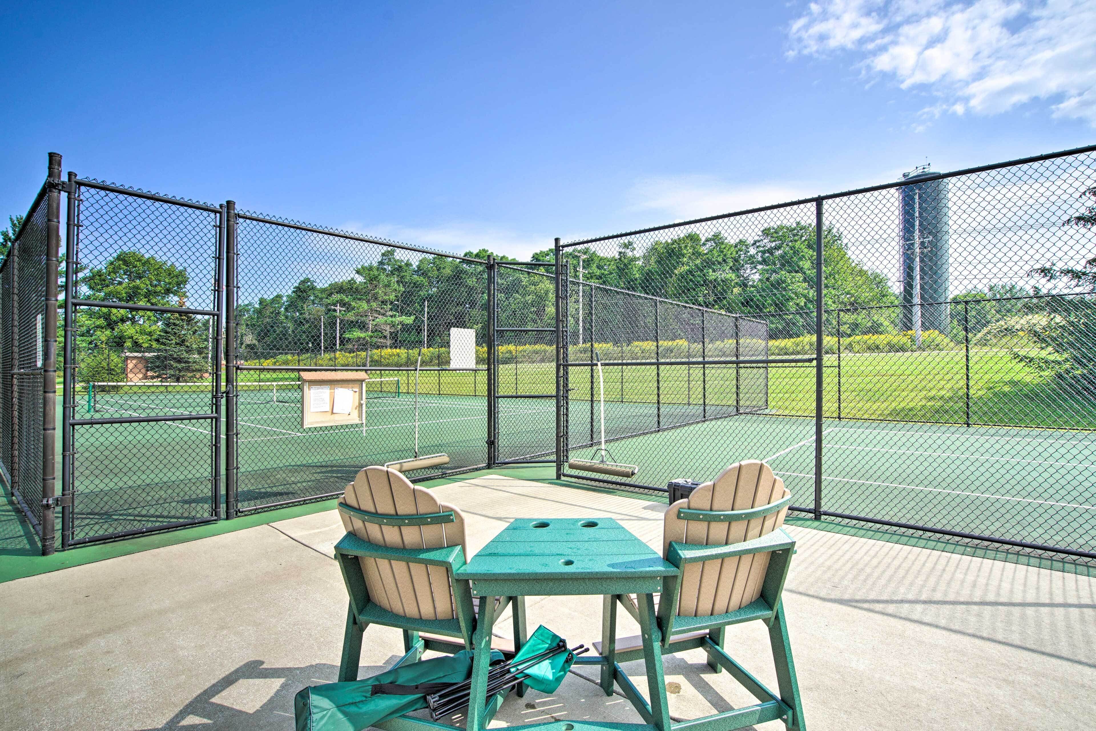 An outdoor tennis court with a seating area, enclosed by a chain-link fence.