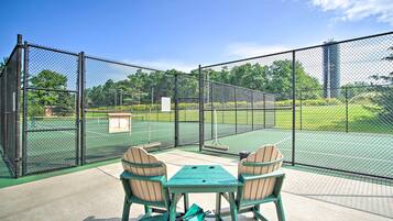 An outdoor tennis court with a seating area, enclosed by a chain-link fence.